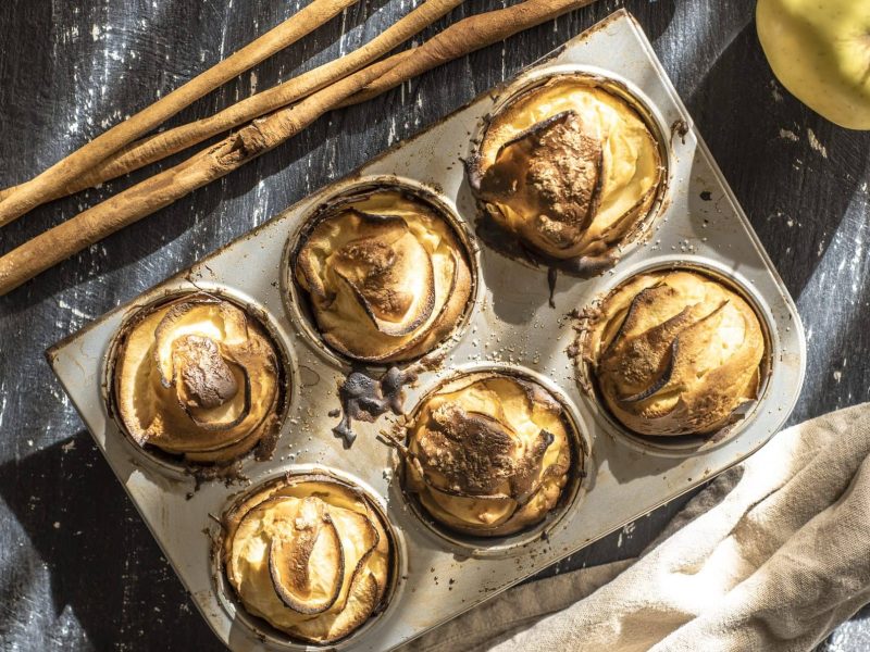 Auf einem dunklen Hintergrund ist eine Muffinform mit sechs saftigen Apfel-Muffins. Dabene liegen Zimtstangen und ein Apfel.