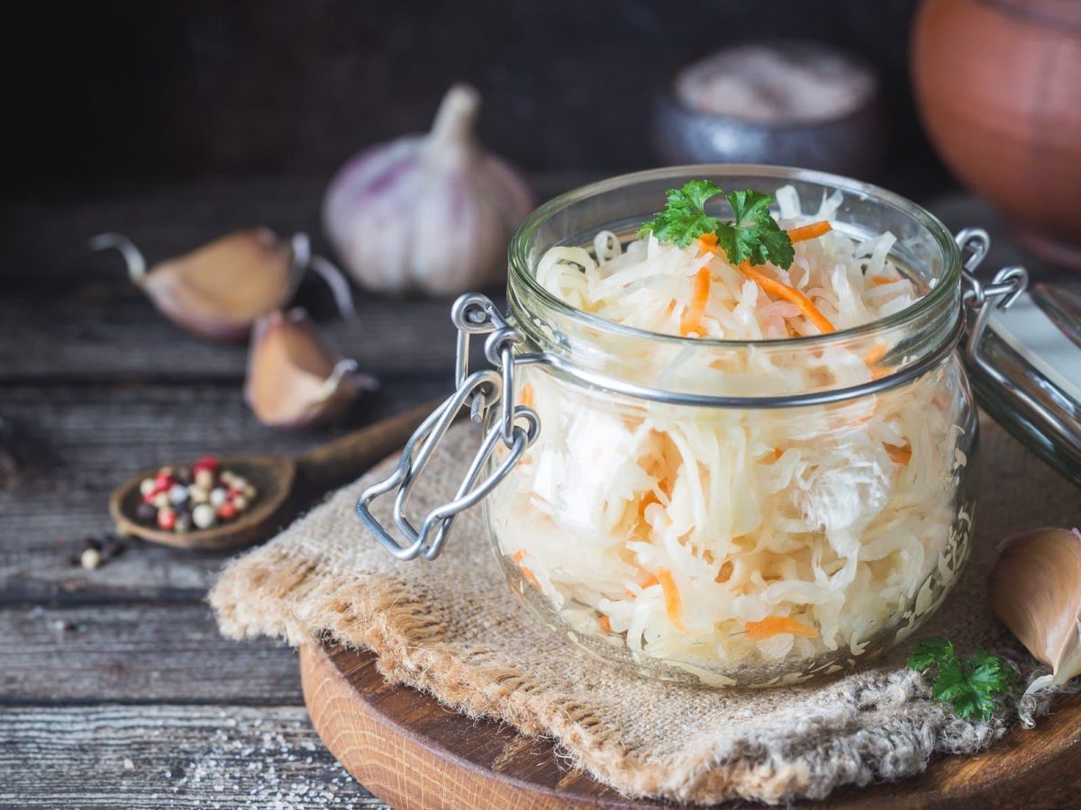 Ein Einmachglas mit Sauerkrautsalat mit Möhren, das auf einem runden Holzbrett steht. Drumherum sieht man Zutaten wie Knoblauch und Pfefferkörner.