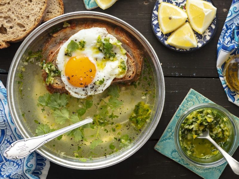 Eine Schüssel Sopa Alentejana neben einer kleinen Glasschüssel mit einer Knoblauch-Koriander-Paste, einer Karaffe mit Olivenöl, einem Teller mit Zitronenstücken und zwei Scheiben Brot, vor einem Holz-Hintergrund, von oben.