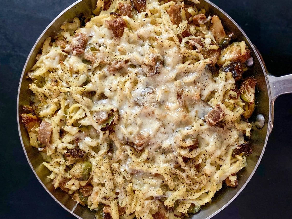 Eine große Portion Spätzle-Rosenkohl-Pfanne mit Bacon und Käse in einer Pfanne präsentiert vor dunklem Hintergrund, von oben.