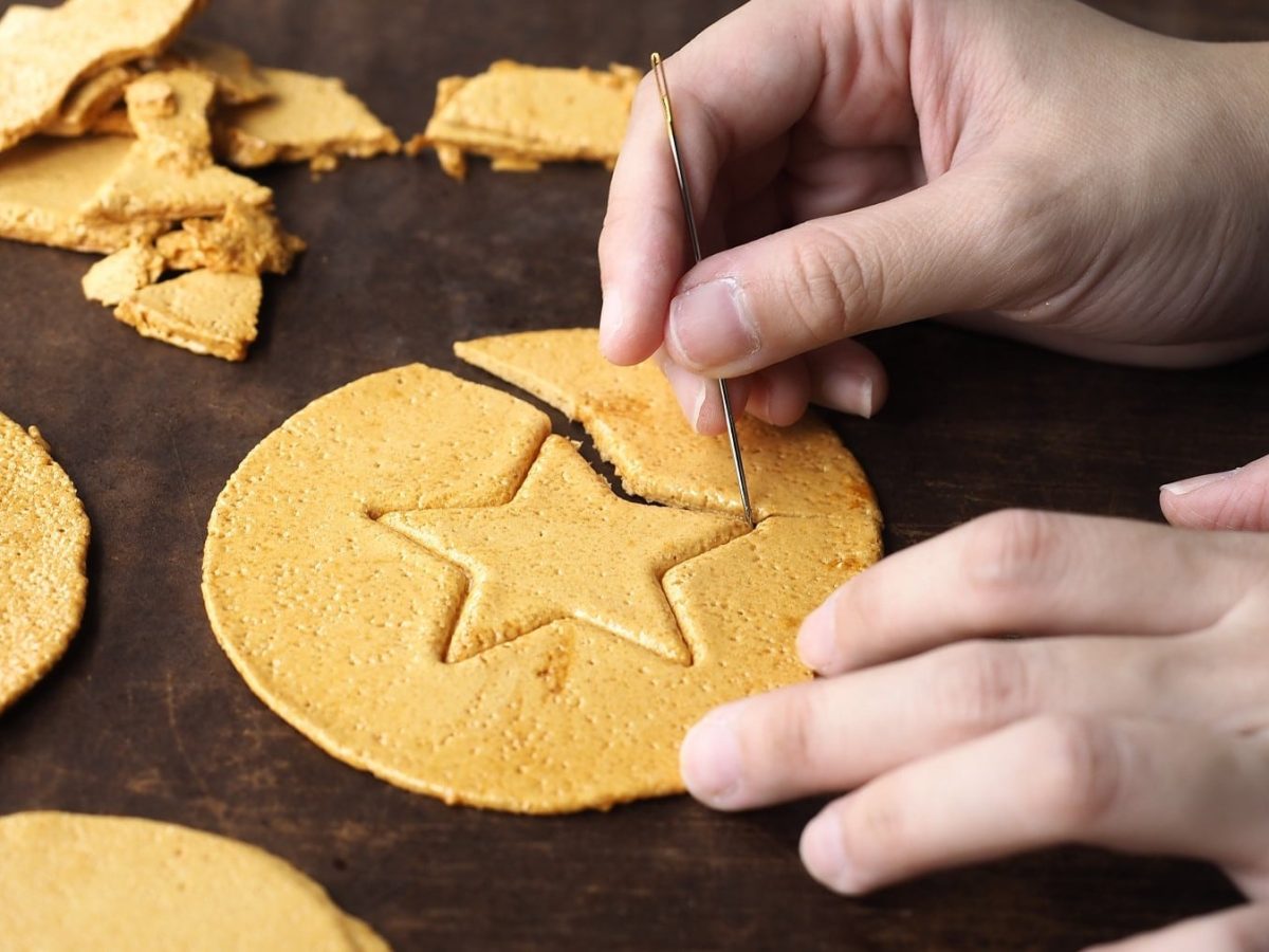 Das "Squid Game" Zuckerspiel Dalgona auf einem braunen Holztisch. Zwei Hände befinden sich auf dem Bild. Eine Hand hält das Zuckerplättchen vorsichtig fest, die andere Hand sticht die Form mit einer Nadel aus.