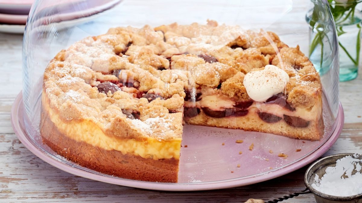 Ein angeschnittener Streuselkuchen mit Pflaumen und Zimt auf einem lila Teller bedeckt mit einer Glas-Glocke neben einer kleinen Schüssel Puderzucker vor einem hellen Hintergrund.