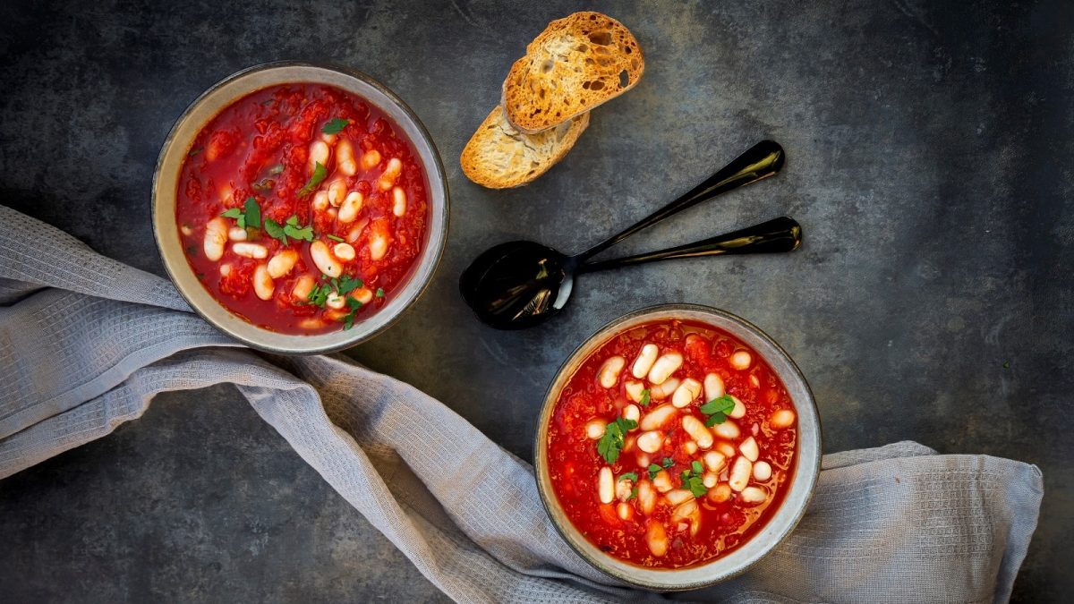 Zwei Schüsseln gefüllt mit jeweils einer Portion der tomatensuppe mit gerösteten Tomaten und Cannellini-Bohnen mit Petersilie garniert, neben zwei Löffeln, zwei Scheiben geröstetem Brot und einem Küchentuch, vor einem dunklen Hintergrund von oben.