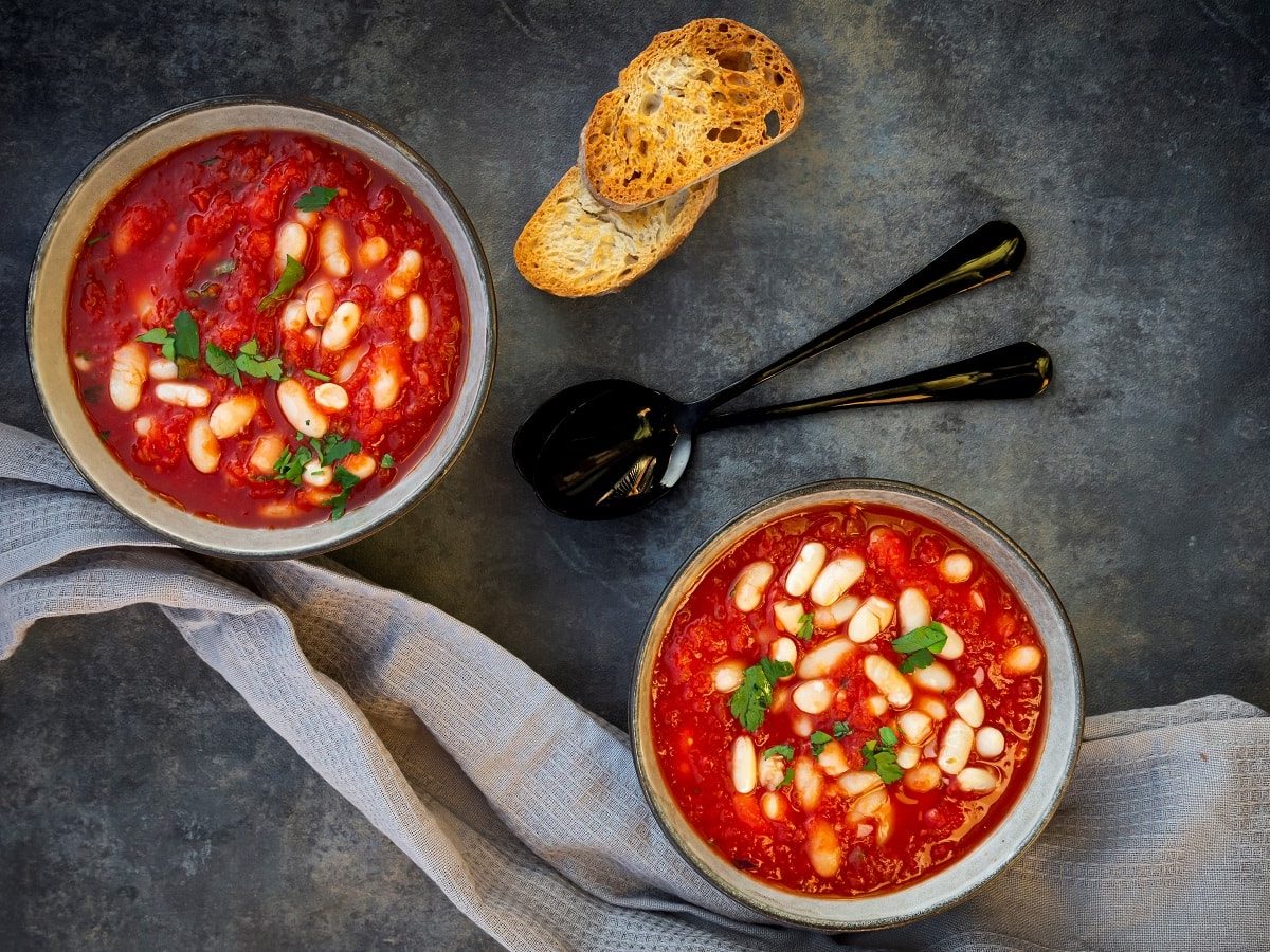 Zwei Schüsseln gefüllt mit jeweils einer Portion der tomatensuppe mit gerösteten Tomaten und Cannellini-Bohnen mit Petersilie garniert, neben zwei Löffeln, zwei Scheiben geröstetem Brot und einem Küchentuch, vor einem dunklen Hintergrund von oben.