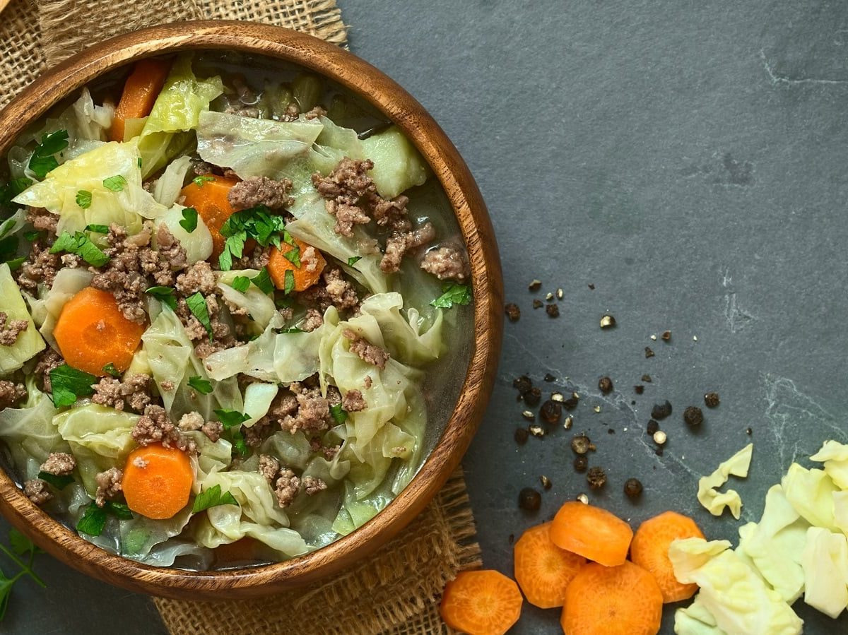 Ein Holzpot mit Weißkohleintopf mit Hackfleisch, Karotten und Kartoffeln, garniert mit Petersilie neben rohen Karotten und Weißkohlstücken, Pfefferkörnern und einem Holzlöffel vor einem grauem Hintergrund, von oben.