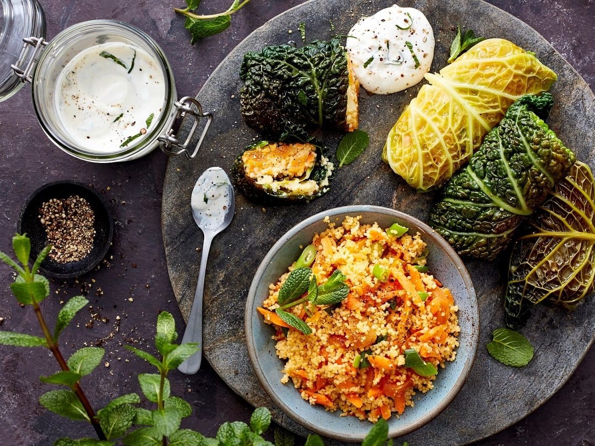 Vier Wirsingrouladen mit Couscous gefüllt auf einer runden Steinplatte zusammen mit einem Klacks Joghurt-Soße und einer Schüssel Gmeüse-Couscous serviert, neben einem Glas mit der Soße, einer kleinen Schüssel mit Pfefferkörnern und frischer Minze, vor einem dunklen Hintergrund, von oben.