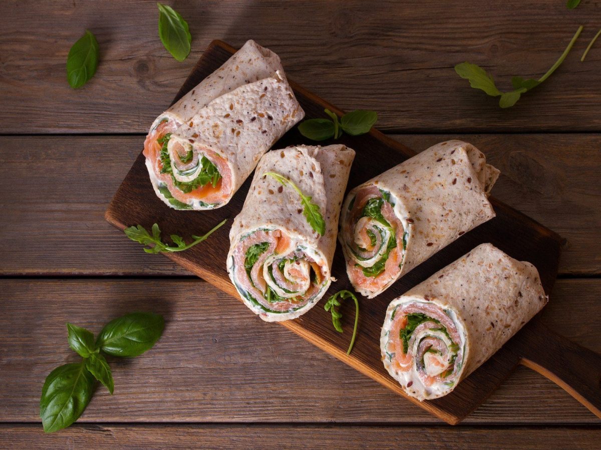 Ein Holzbrett auf einem Holz-Untergrund, auf dem Wraps mit Lachs und Meerrettich liegen. Drumherum ein paar Rucolablätter. Alles in der Draufsicht.