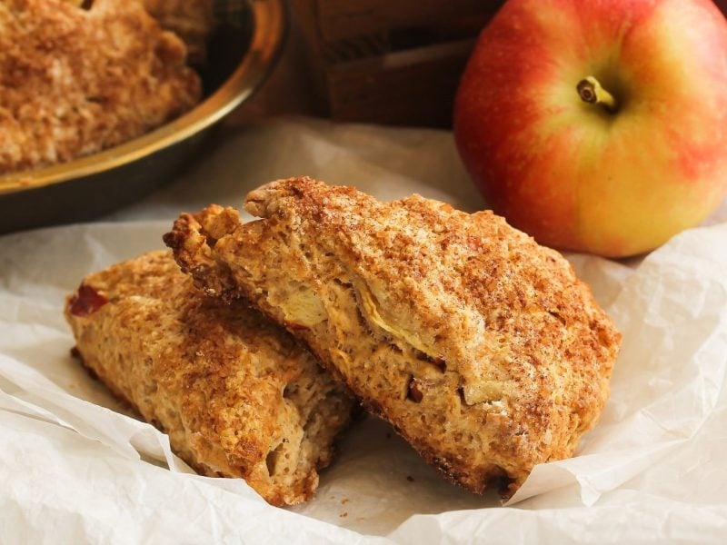 Zwei Apfel-Scones liegen auf gestapelt übereinander auf hellem Backpapier. Im Hintergrund liegt ein Apfel und eine Schüssel mit Scones.