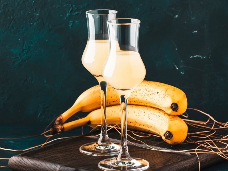 Zwei Gläser mit Bananen-Likör stehen auf einem Holzbrett auf einem dunklen Untergrund. Der Hintergrund ist aus blau-schwarzen Steinen. Hinter den Gläsern liegen zwei Bananen und einige trockene Gräser.