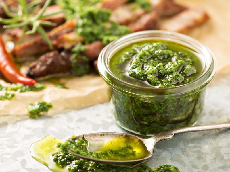 Veganes grünes Pesto in Glas auf Serviette au der Frontalansicht. Davor ein Teelöffel mit Pesto und Olivenöl, das leicht verläuft. Im Hintergrund Kräuter, Chili und Nüsse leicht verschwommen auf Holzbrett angedeutet.