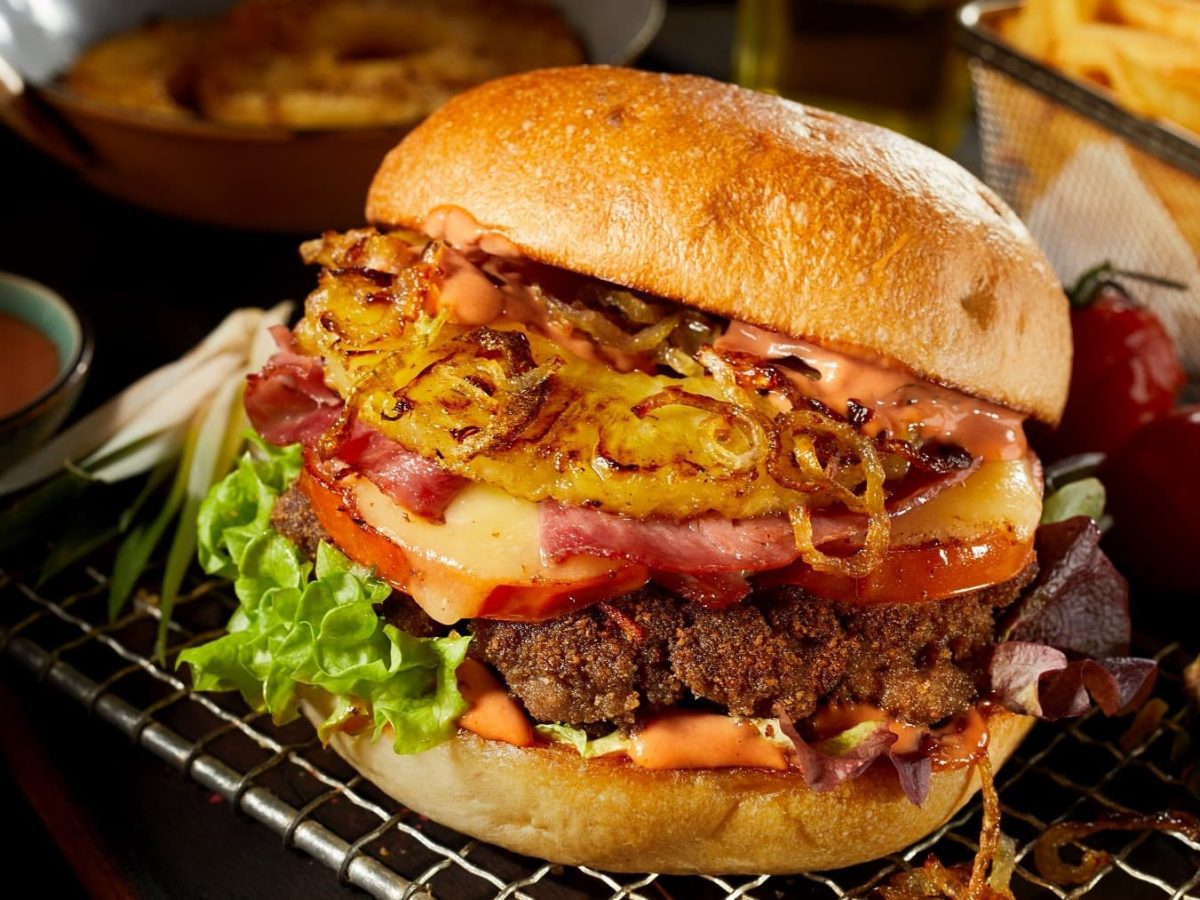 Ein Burger-Hawaii mit Ananas, Speck und gebratenen Zwiebeln liegt auf einem Gitter. Um den Burger herum steht eine Schale mit Coktailsuppe, Ananasstückchen und einige Tomaten. Im Hintergrund liegen weitere Ananasscheiben und eine Portion Pommes.