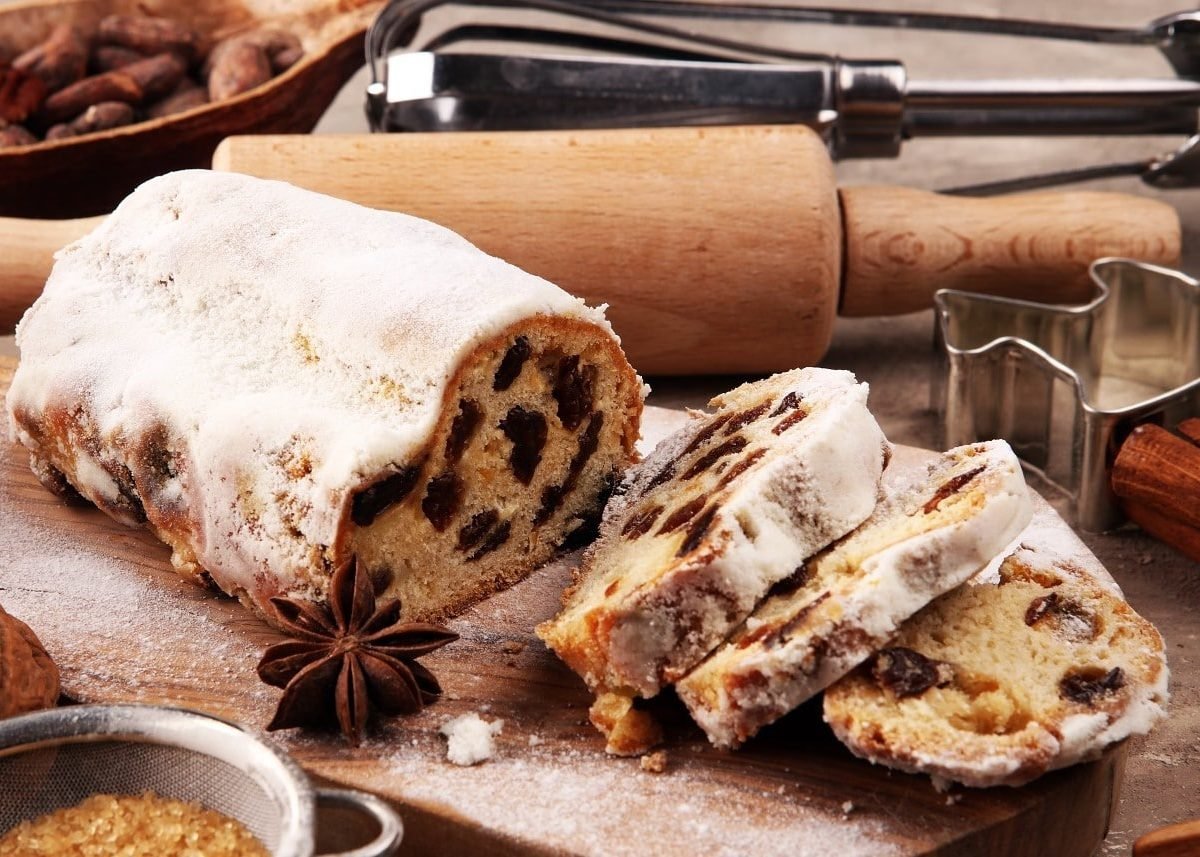 Ein angeschnittener Christstollen steht auf einem Holzbrett umgeben von verschiedenen Backutensilien, Nüssen, Zucker und Zimtstangen. Der Stollen ist verziert mit Puderzucker und angerichtet mit Sternanis.