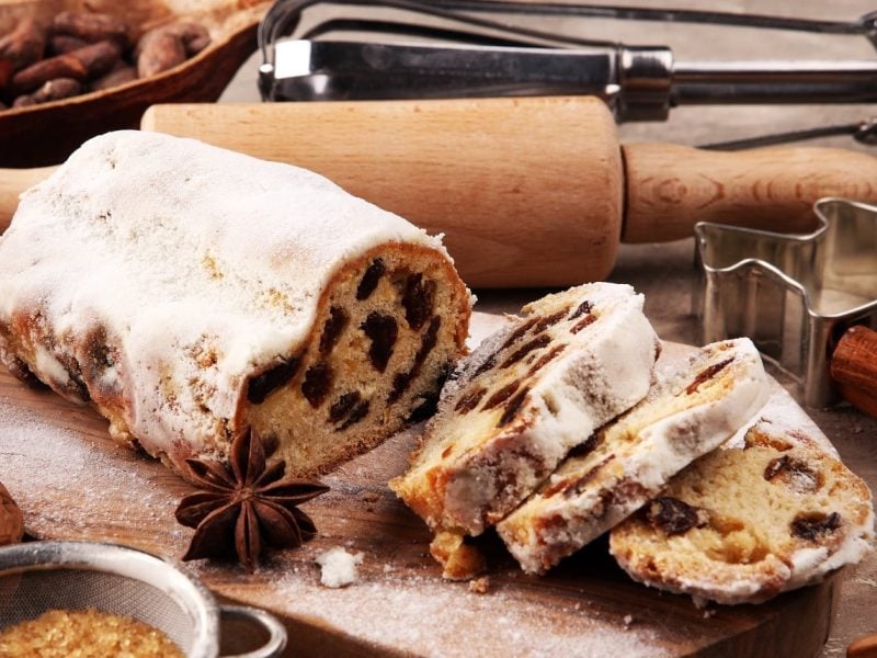 Ein angeschnittener Christstollen steht auf einem Holzbrett umgeben von verschiedenen Backutensilien, Nüssen, Zucker und Zimtstangen. Der Stollen ist verziert mit Puderzucker und angerichtet mit Sternanis.