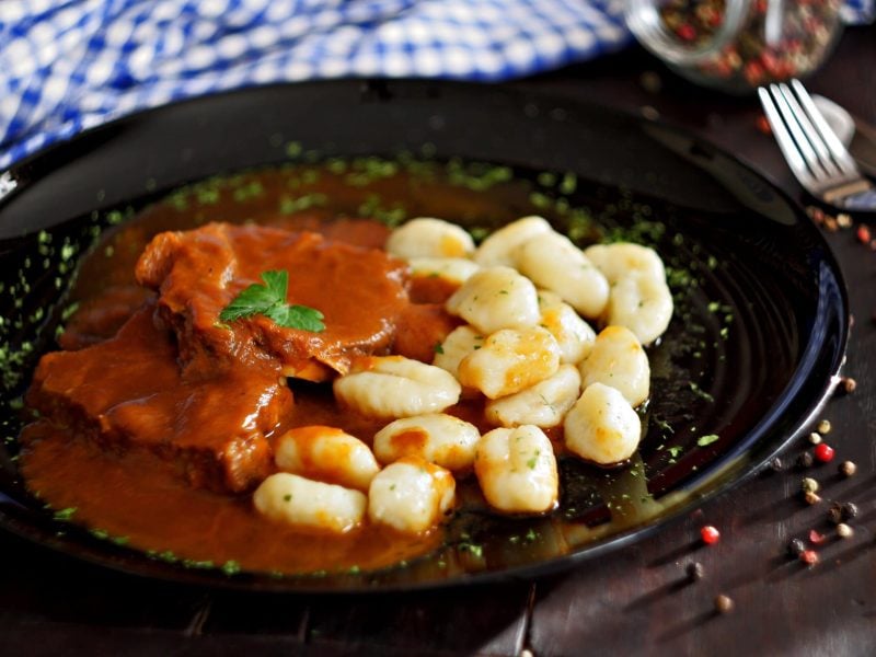 Der Dalmatinische Rinderbraten angerichtet mit Kartoffelgnocchi auf einem schwarzen Teller. Dazu Pfefferkörner als Deko.