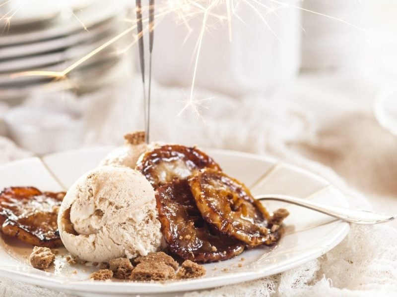 Unser Spekulatius-Eis weihnachtlich mit Wunderkerze auf weißen Tellern und Geschirr im Hintergrund in Szene gesetzt.