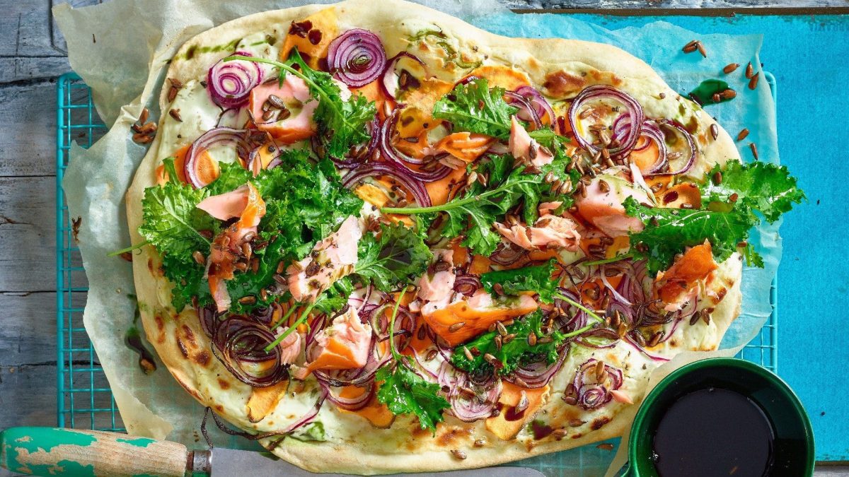 Unser Flammkuchen mit Grünkohl und Lachs liegt ausgebreitet auf einem türkisfarbenen Gitter mit Backpapier.