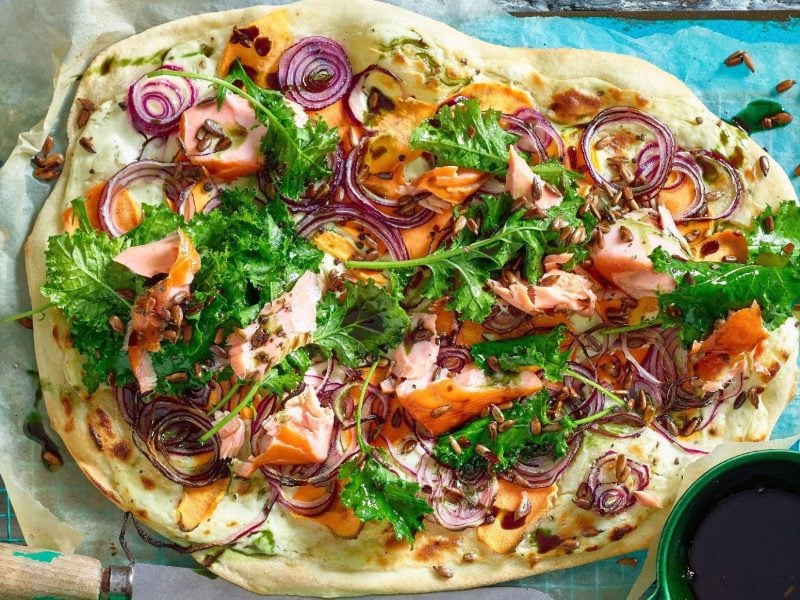 Unser Flammkuchen mit Grünkohl und Lachs liegt ausgebreitet auf einem türkisfarbenen Gitter mit Backpapier.