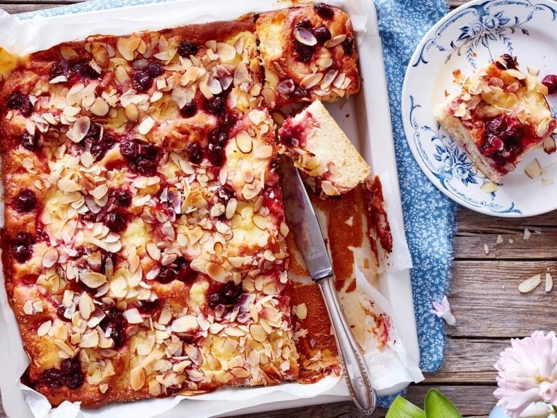 Ein Kiba-Pudding-Butterkuchen in einer Form steht auf einem blau gemusterten Geschirrtuch auf einem braunen Holztisch. Der Kuchen ist bereits angeschnitten und ein Stück steht auf einem blau-weißen Teller mit Blütenmuster. In der Form liegt ein Messer. Rechts neben dem Kuchen steht eine kleine Schüssel mit Mandelblättchen.