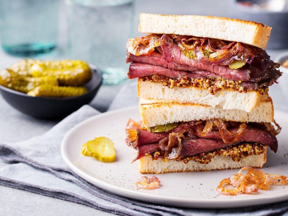 Pastrami-Sandwich mit Senf, karamellisierten Zwiebeln und Gurken auf einem weißen Teller, umgeben von Zutaten.