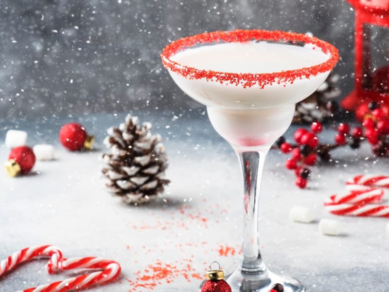 Ein Schneeflocken-Daiquiri in einem mit Zuckerperlen verzierten Glas auf einem Untergrund aus Stein. Um das Glas herum liegen Zuckerstangen, Kienäpfel und Christbaumkugeln. Künstlicher Schnee rieselt im Hintergrund.