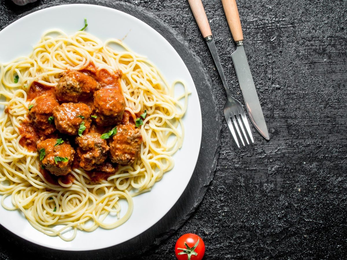 Spaghetti mit Fleischbällchen aus "Der Pate" auf weißem Teller und schwarzem Tisch aus der Vogelperspektive. Hackbällchen in Tomatensoße auf Spaghetti. Rechter Bildrand Messer und Gabel mit Holzgriff. Einzelne Tomate am unteren Bildrand zu sehen.