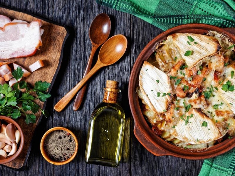 Tartiflette mit Reblochon und Speck in Auflaufform auf dunklem Holz, umgeben von verschiedenen Zutaten.