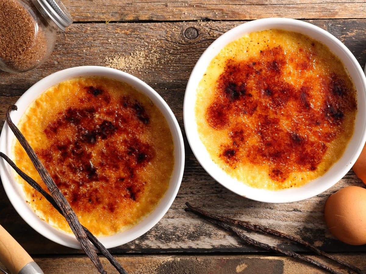 Zwei weiße Förmchen mit Amelies Crème Brûlée mit extra krosser Karamellschicht gefüllt, neben Vanilleschoten, Eiern, Zucker und Milch, vor einem Holzhintergrund, von oben.