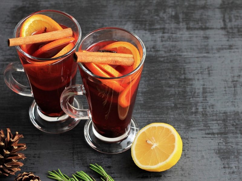 Zwei hohe Gläser gefüllt mit dem Apfel-Holunder-Orangen Punsch mit viel Gewürzen, garniert mit Orangenscheiben und Zimtstangen, neben einer halben Zitrone, einem Tannenzapfen und einem Tannenzweig.