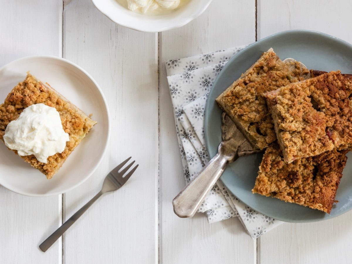 In der Draufsicht sieht man zwei Teller mit einigen Stücken Apfel-Schmand-Kuchen auf weißem Holz. Außerdem Servietten und eine Kuchengabel.