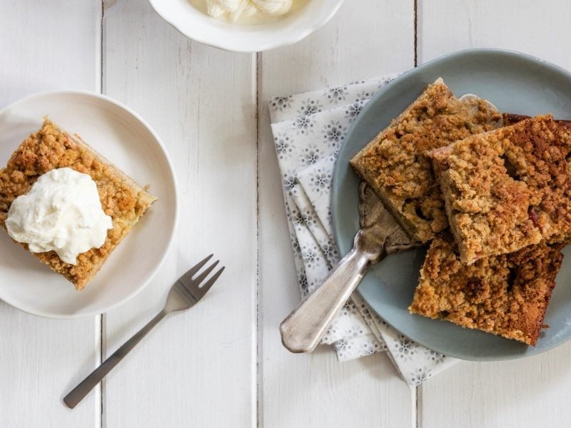 In der Draufsicht sieht man zwei Teller mit einigen Stücken Apfel-Schmand-Kuchen auf weißem Holz. Außerdem Servietten und eine Kuchengabel.