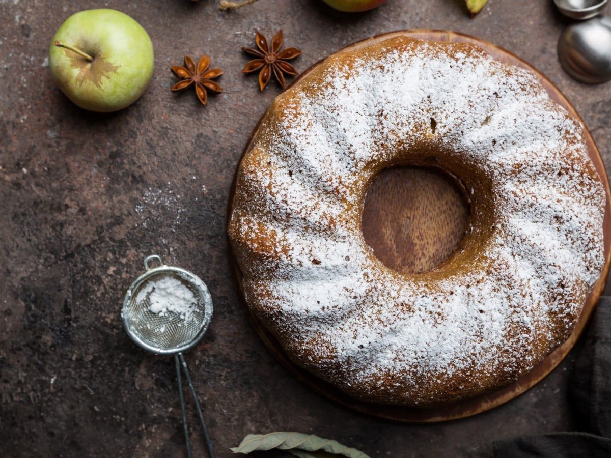 Ein Apfelkuchen mit Kardamom, ein Topfkuchen, in der Draufsicht. Daneben liegen Äpfel, ein Sieb mit Puderzucker und Sternanis.