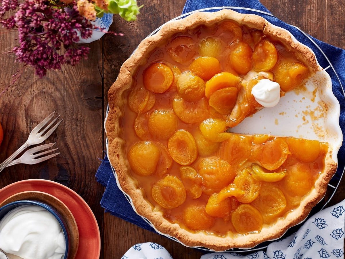 Eine Tarte-Form gefüllt mit der Aprikosen-Tarte mit Espresso-Creme bei der ein Stück fehlt und die mit etwas Schlagsahne garniert ist, neben einer Schüssel Schlagsahne, einem Glas Konfitüre, Gabeln und Blumen vor einem Holzhintergrund, von oben.