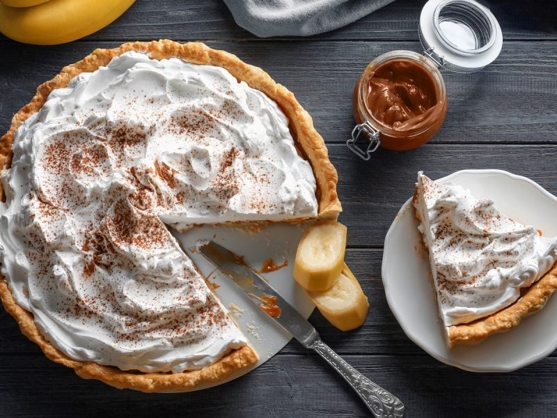 Ein angeschnittener Banoffee Pie wie aus dem Film Tatsächlich Liebe neben einem Messer, Bananenscheiben, einem Glas mit Karamell und einem Teller mit einem Stück des Kuchens vor einem grauen Hintergrund von oben.