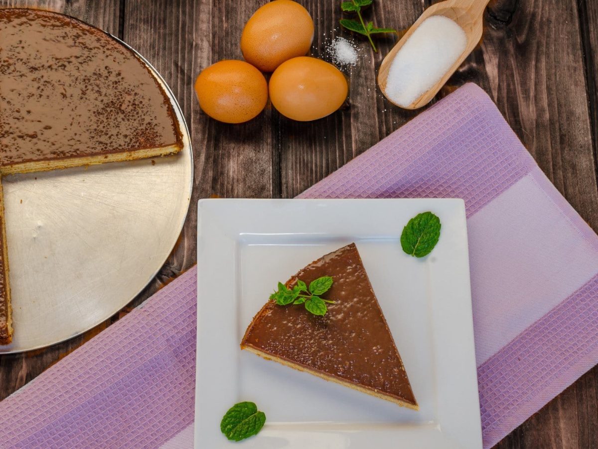 Ein angeschnittener Baumkuchen auf rustikalem Holzuntergrund in der Draufsicht. Außerdem Eier und ein Holzlöffel Zucker.