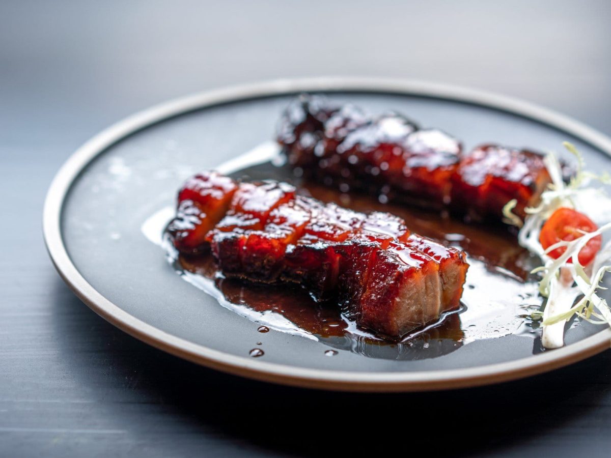 Aufgeschnittenes Schweinefleisch Char Siu aus der Frontalsicht. Rotbräunliche Färbung durch Röstung auf grauem Teller und grauem Untergrund. Rechts aufgeschnittener weißer Rettich..