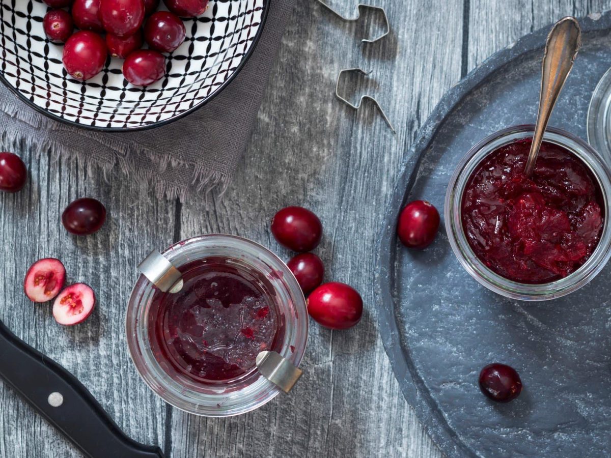 In der Draufsicht sieht man Weckgläser mit Cranberry-Soße, außerdem frische Cranberries und ein Messer auf Holzplanken.