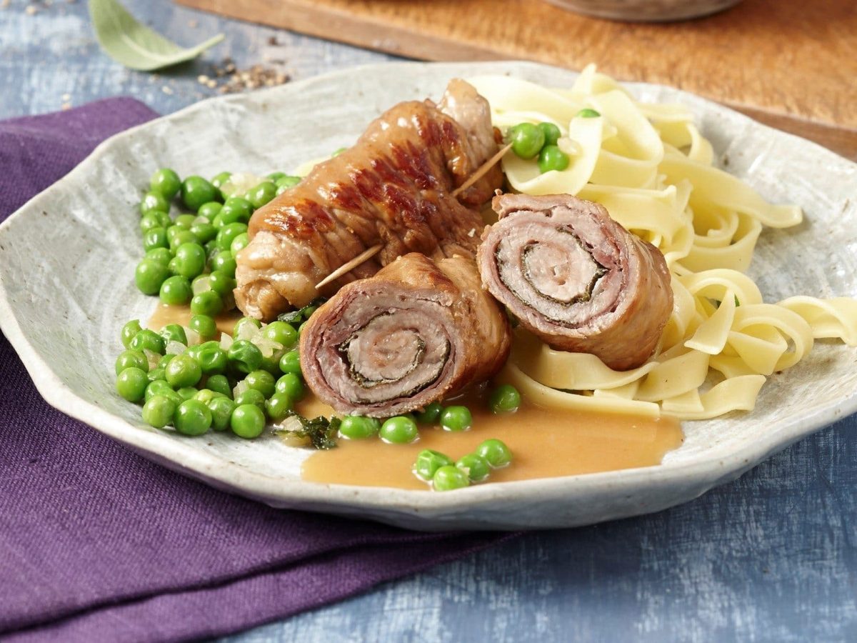 Auf einem Teller liegt eine Roulade. Sie ist aus zartem Kalbsfleisch und wird mit Salbei gewürzt. Dazu gibt es Erbsen und Nudeln