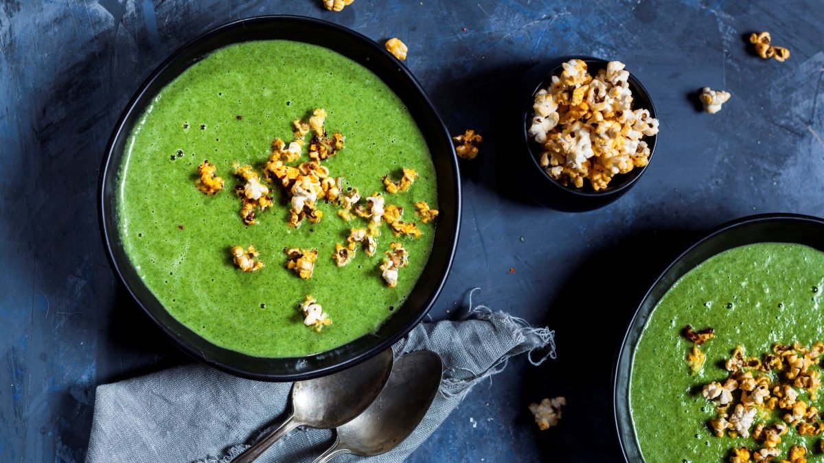 Zwei Schüsseln Erbsen-Spinat-Suppe mit Popcorn in der Draufsicht, daneben steht eine kleine Schale mit Popcorn, außerdem liegen zwei Löffel bereit.