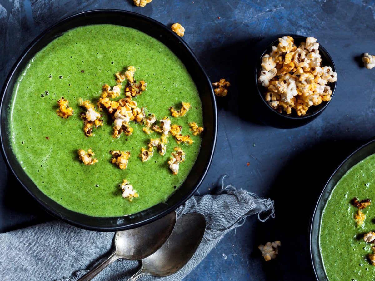 Zwei Schüsseln Erbsen-Spinat-Suppe mit Popcorn in der Draufsicht, daneben steht eine kleine Schale mit Popcorn, außerdem liegen zwei Löffel bereit.