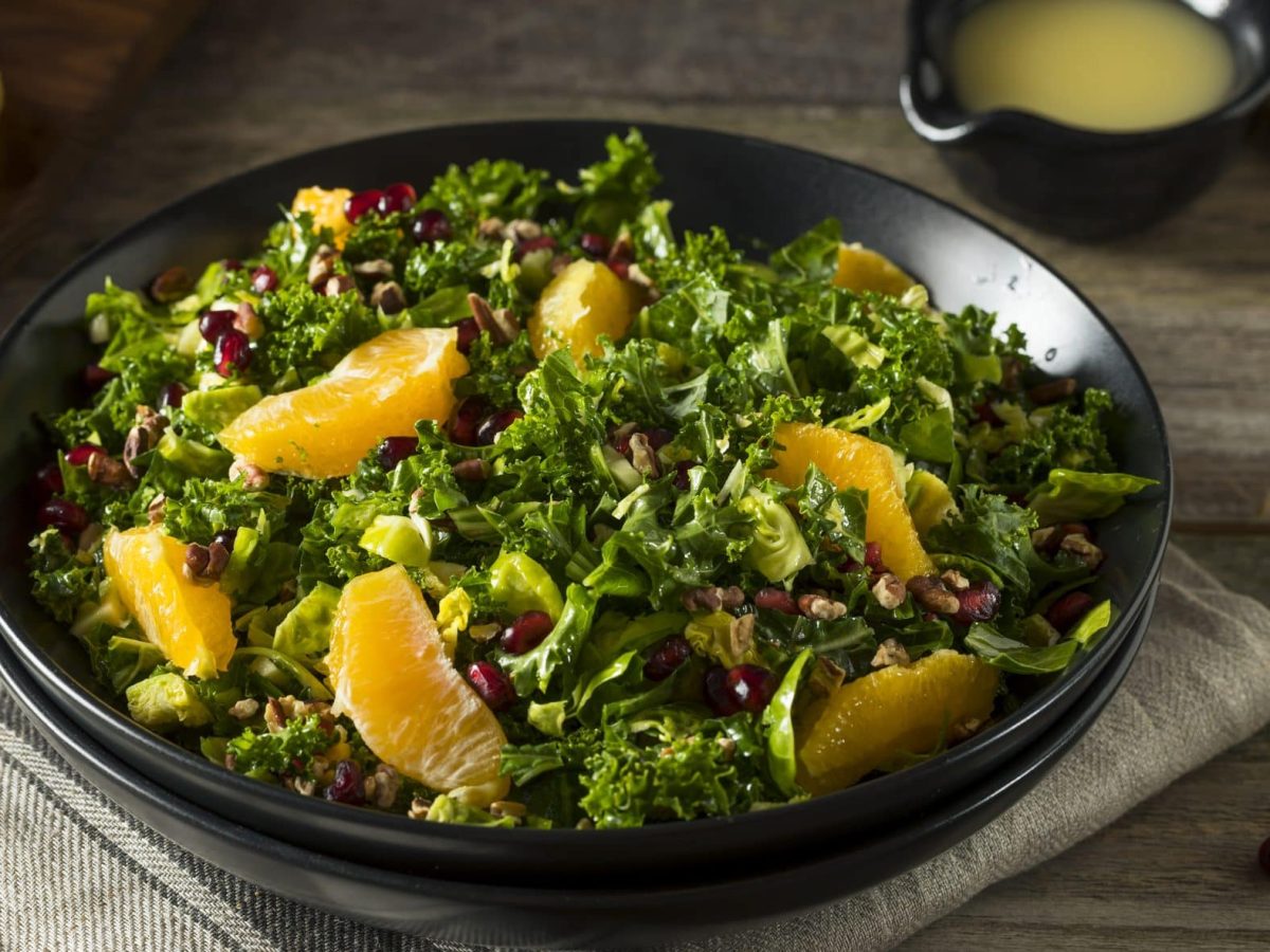 In einer runden Schale ist Grünkohlsalat mit Orange und Walnüssen Auf dem Tisch sind zwei Orangenhälften zu sehen.