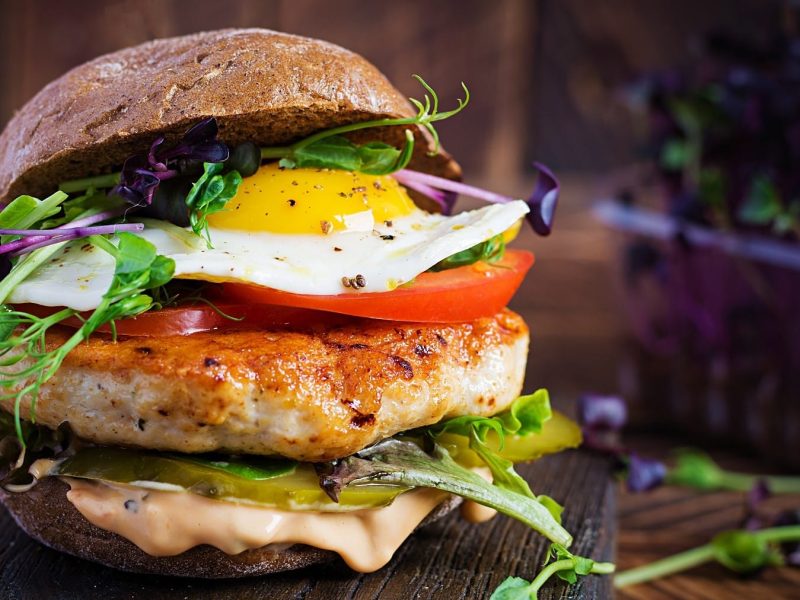 Ein Hähnchen-Burger mit Spiegelei steht auf einem dunklen Holzbrett auf einem dunklen Untergrund. Im Hintergrund steht eine Schale mit Salat.