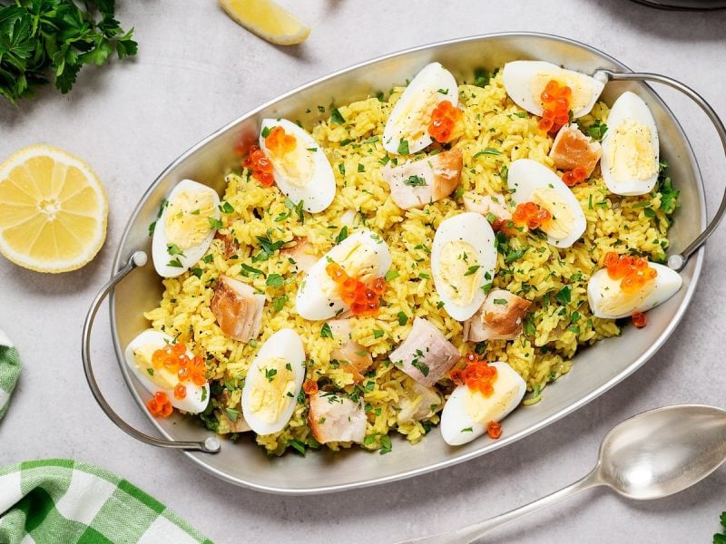 Eine Servier-Schüssel angerichtet mit Kedgeree, einem indischen Gericht mit britischer Adaption, garniert mit rotem Kaviar und Petersilie, neben einem Löffel, einer halben Zitrone, Salz, Pfeffer, Petersilie und einem Tuch, vor grauem Hintergrund, von oben.