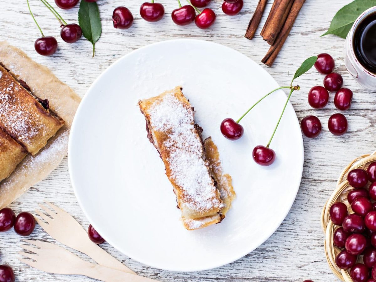 Draufsicht von oben: Auf einem weißen Tisch liegen Kirschen. Auf einem weißen Teller liegen mehrere Stücke Kirschstrudel.