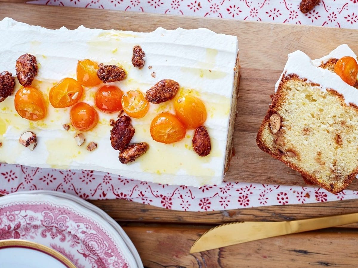 Ein Holzbrett auf dem der Kuchen mit gebrannten mandeln und Kumquats liegt, angeschnitten mit zwei Stücken daneben