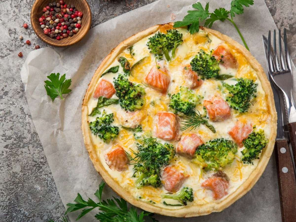 In der Draufsicht: Eine Brokkoli-Lachs Quiche ist in einer runden Form. Daneben liegen Messer und Gabel. Auch eine Schale mit Pfefferkörnern ist zu sehen.