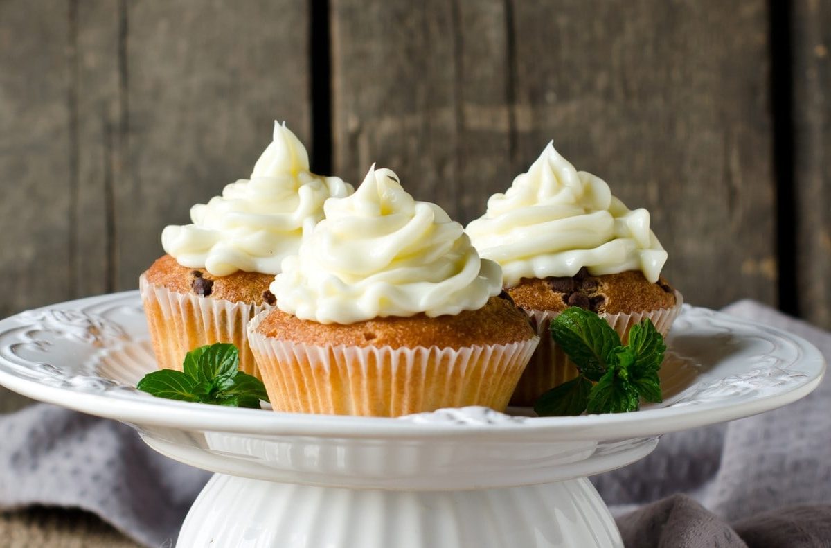 Auf einem Kuchenständer sehen drei Macadamia-Cupcakes mit Frosting vor einer Holzwand.