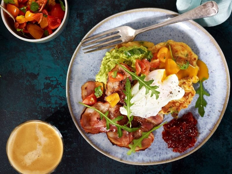 Ein Teller mit den Maispfannkuchen mit Bacon und pochiertem Ei mit Avocado-Creme bestrichen und Rucola und Kirchtomaten garniert, neben einer Tasse Kaffee, einer Schüssel Rucola und einer kleinen Schale Kirschtomaten vor einem dunkelgrünen Hintergrund, von oben.