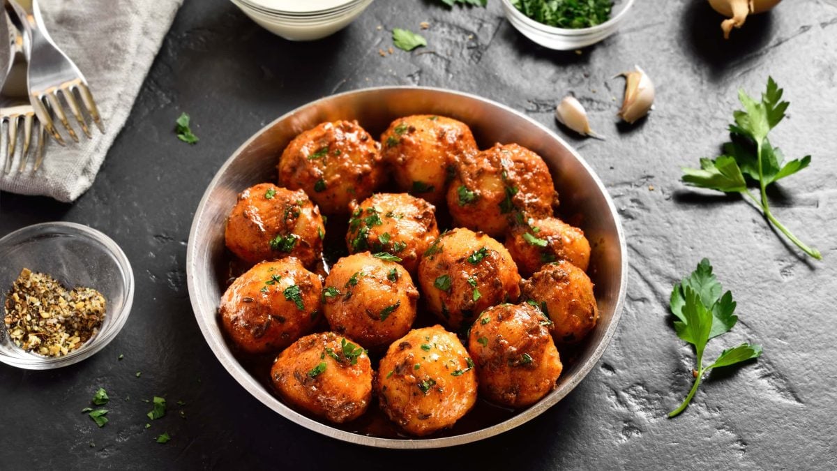 Eine Schale mit Bombay-Kartoffeln, Masala Aloo, auf dunklem Untergrund. Drumherum liegen Zutaten wie Knoblauch oder Gewürze, außerdem zwei Gabeln.