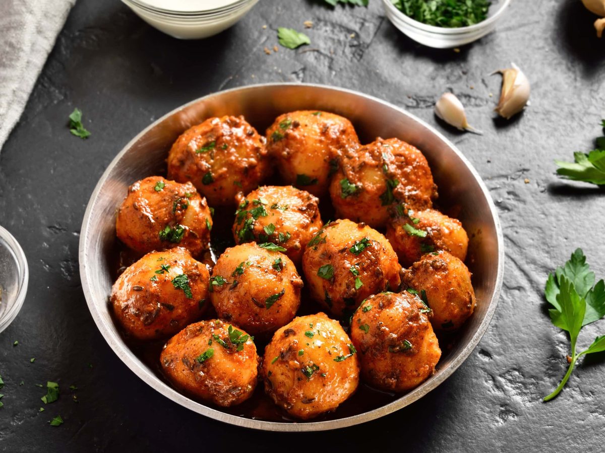 Eine Schale mit Bombay-Kartoffeln, Masala Aloo, auf dunklem Untergrund. Drumherum liegen Zutaten wie Knoblauch oder Gewürze, außerdem zwei Gabeln.