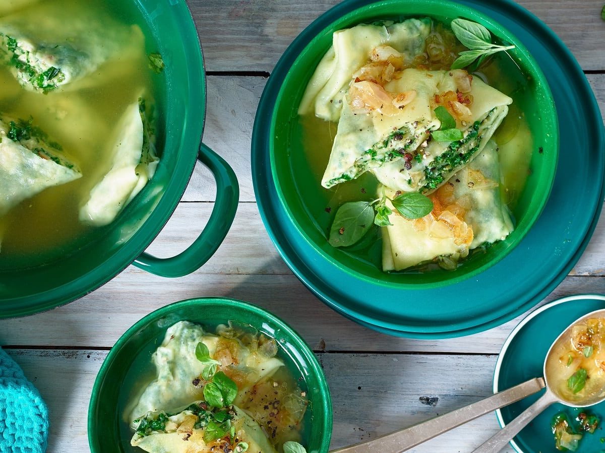 Ein grüner Topf und zwei kleinere Schüsseln mit Portionen der Maultaschen mit Tofu und Grünkohl neben einem kleinen Teller mit Löffeln und einem blauen gehäckelten Untersetzter, vor einem Holzhintergrund, von oben.